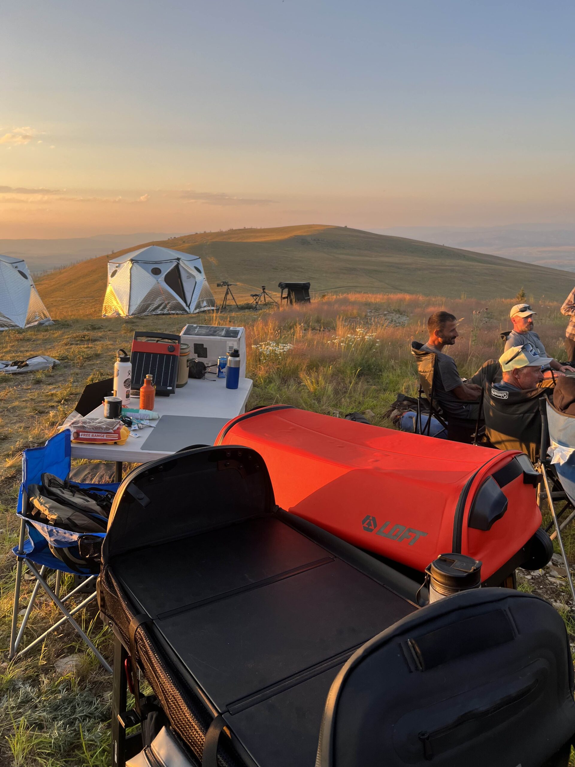 Two duffle bags in the foreground of a camping setting with seated people in the background.